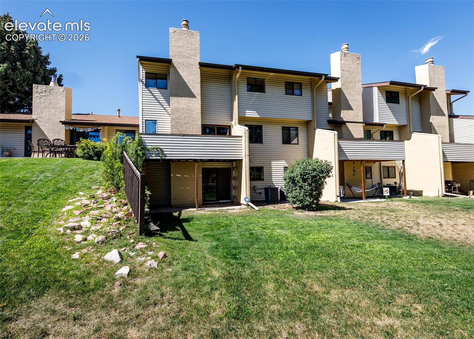 Multi-level exterior featuring horizontal siding, exterior chimneys, private ground-level patios, and a shared open lawn area with rock landscaping