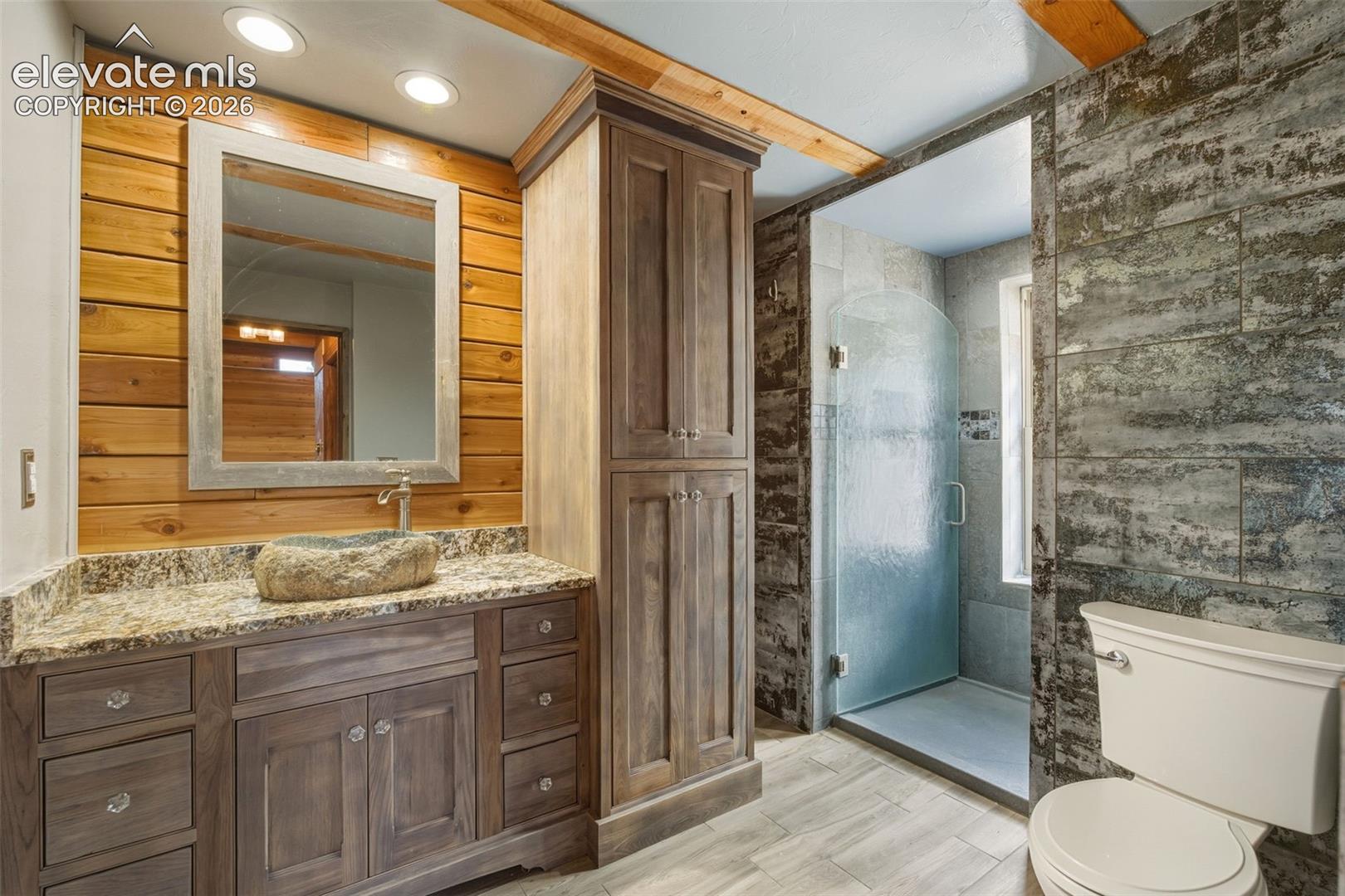 Wood-paneled wall with recessed lighting, stone vessel sink, granite countertop, wood-finish vanity with storage, and a glass-enclosed shower
