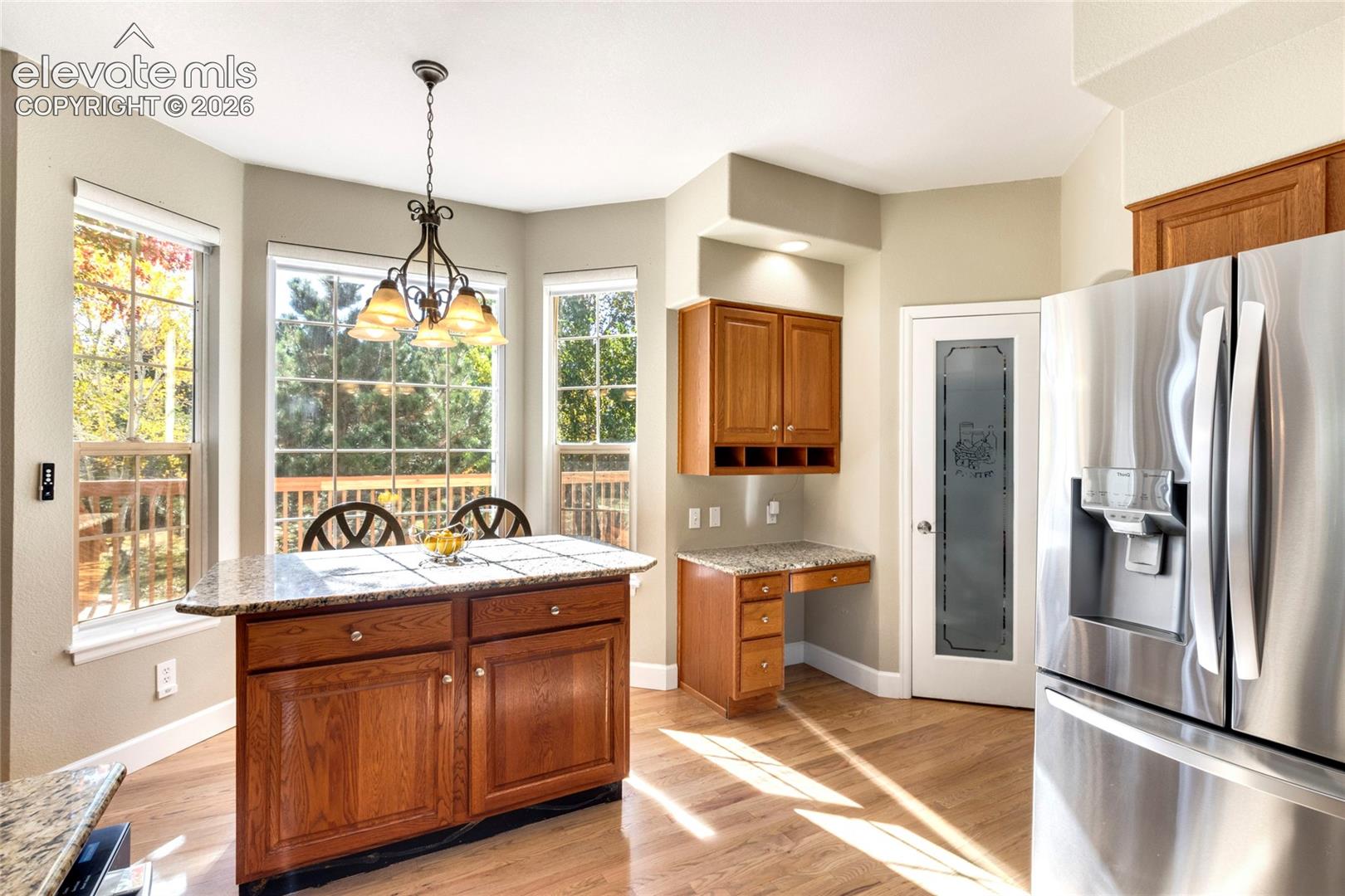 Kitchen featuring a wood-finish island with stone countertop, built-in desk cabinetry, stainless steel refrigerator, paneled-glass pantry door, and bay window surround with metal-finish chandelier