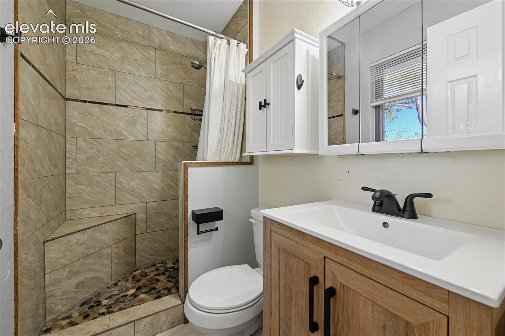 Primary Bathroom featuring a walk-in shower with stone-tile surround, integrated corner bench, and river-rock flooring, paired with a wood-finish vanity topped with a rectangular sink and matte black fixtures