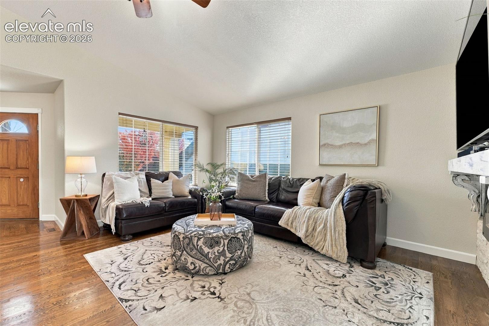Living area featuring vaulted ceilings, wood-finish flooring, large windows with blinds, and a natural stone fireplace surround