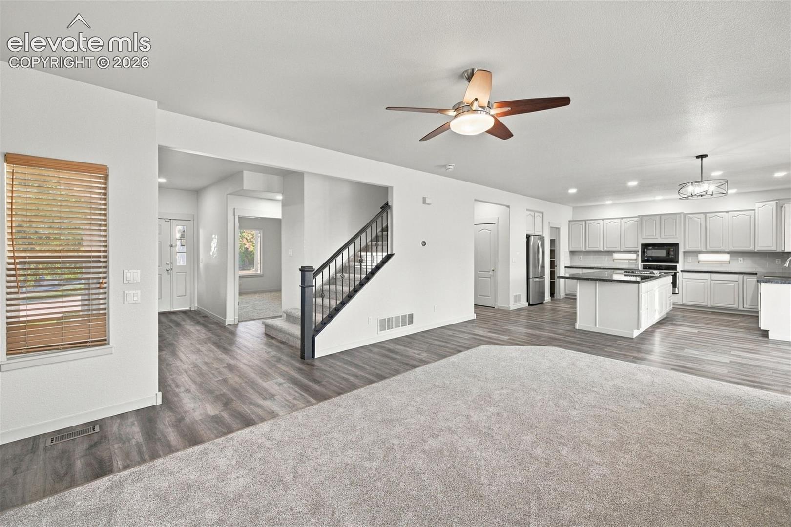 Open-concept floor plan featuring wood-finish flooring, a kitchen with white cabinetry and a center island, recessed lighting, and a staircase with a dark metal railing