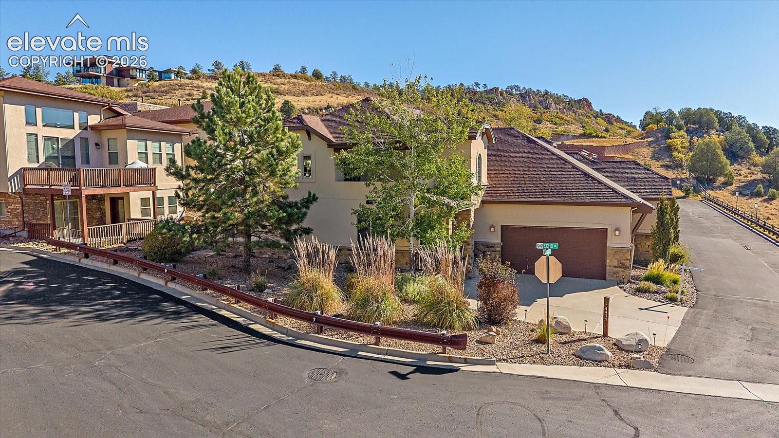 Multi-story stucco and stone exterior featuring a shingled roof, attached multi-car garage, elevated wooden deck, and landscaped grounds with mature trees