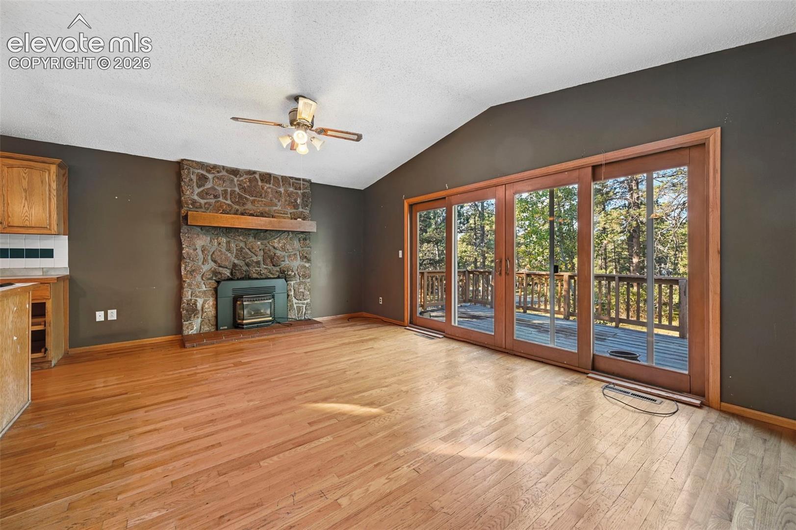 Vaulted ceiling with a ceiling fan, stone-clad fireplace with wood mantle, wide plank wood-finish flooring, and sliding glass doors opening to a wood deck