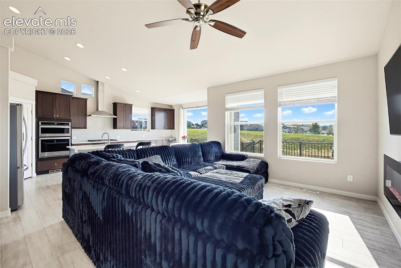 Open-concept floor plan featuring vaulted ceilings, recessed lighting, wood-finish flooring, dual wall-mounted ovens, and large windows with landscape scenery