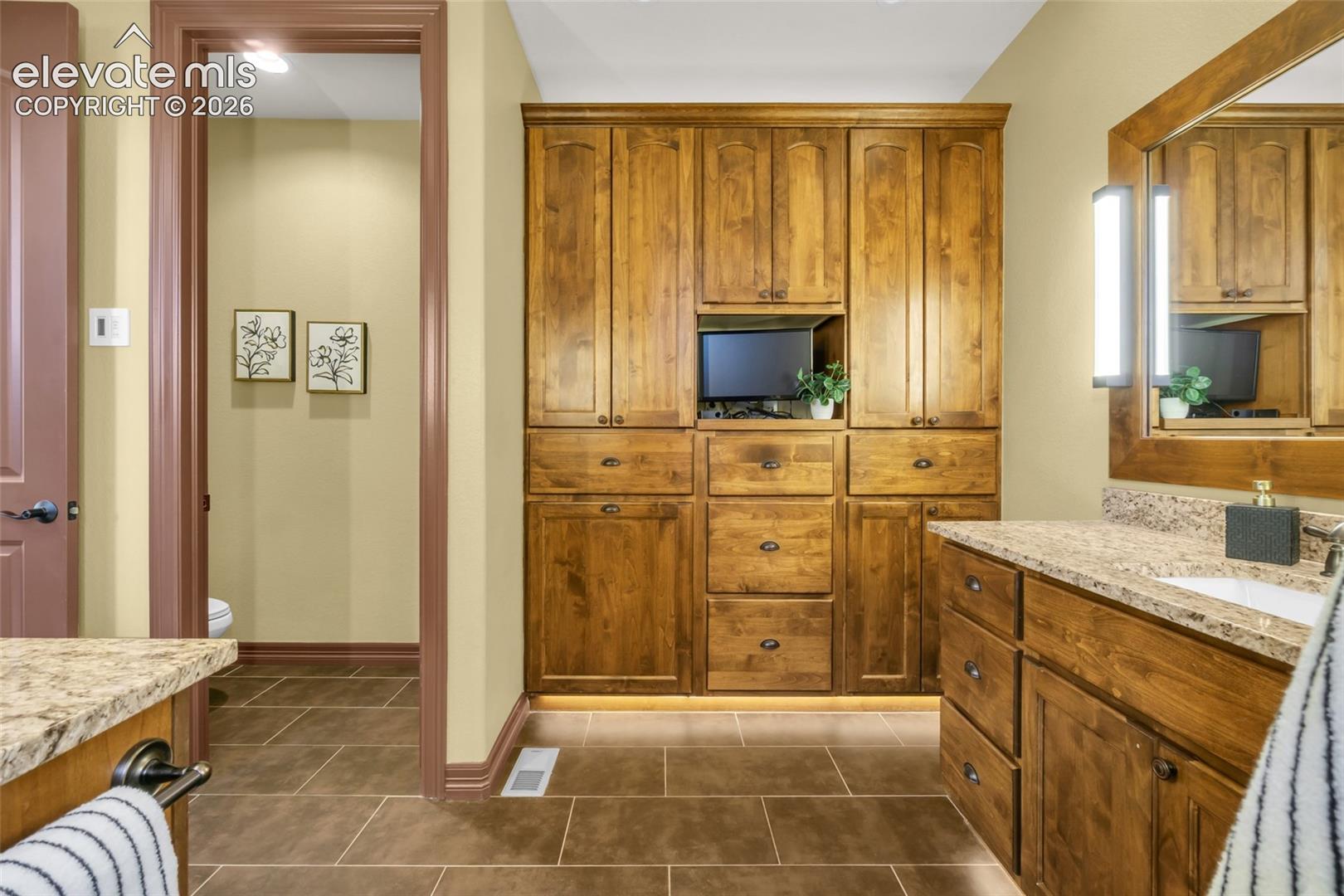 Custom built-in wood cabinetry, stone-finish countertops, tiled flooring, wood-framed mirror with integrated lighting, and crown molding