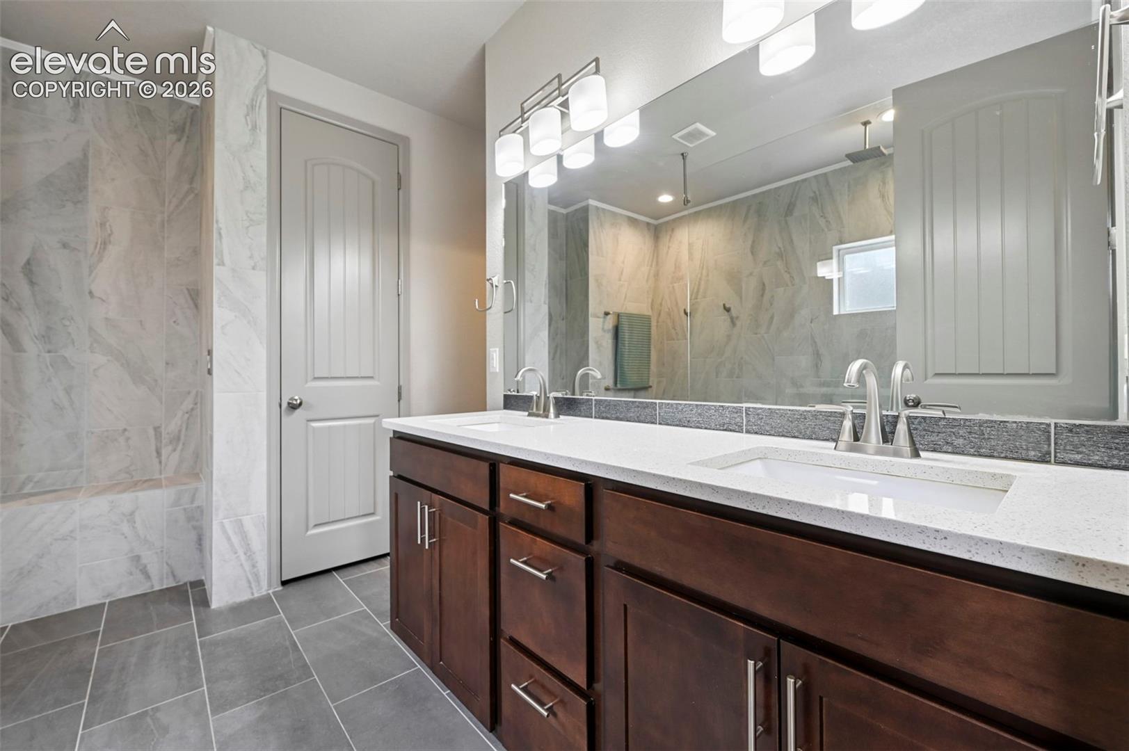 Double vanity with dark wood-finish cabinetry, quartz countertops, undermount sinks, and a full-width mirror with multi-bulb lighting, complemented by tile flooring and a walk-in tiled shower