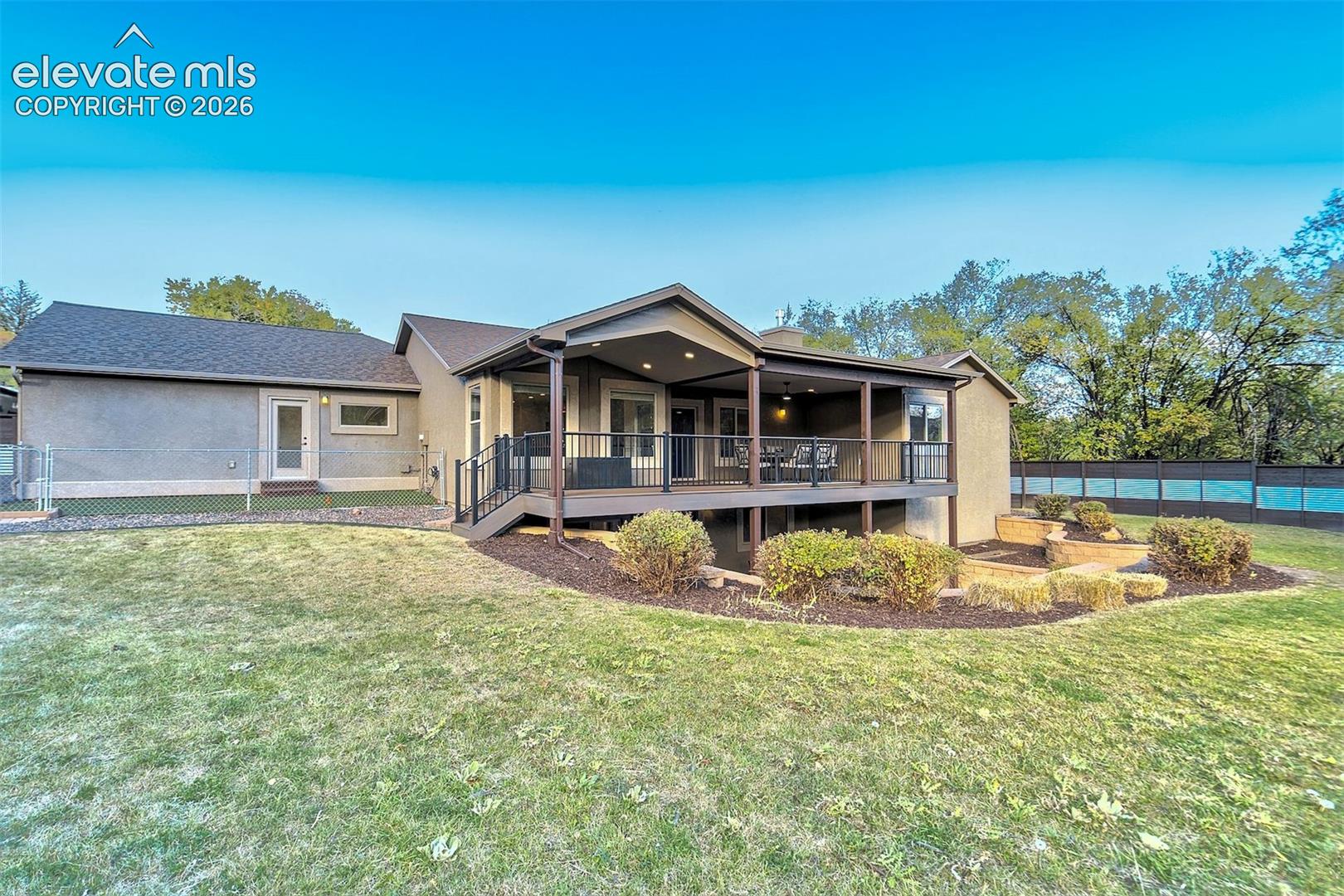 Stucco exterior featuring a covered rear deck with metal railing, tiered landscaping, and a fenced yard