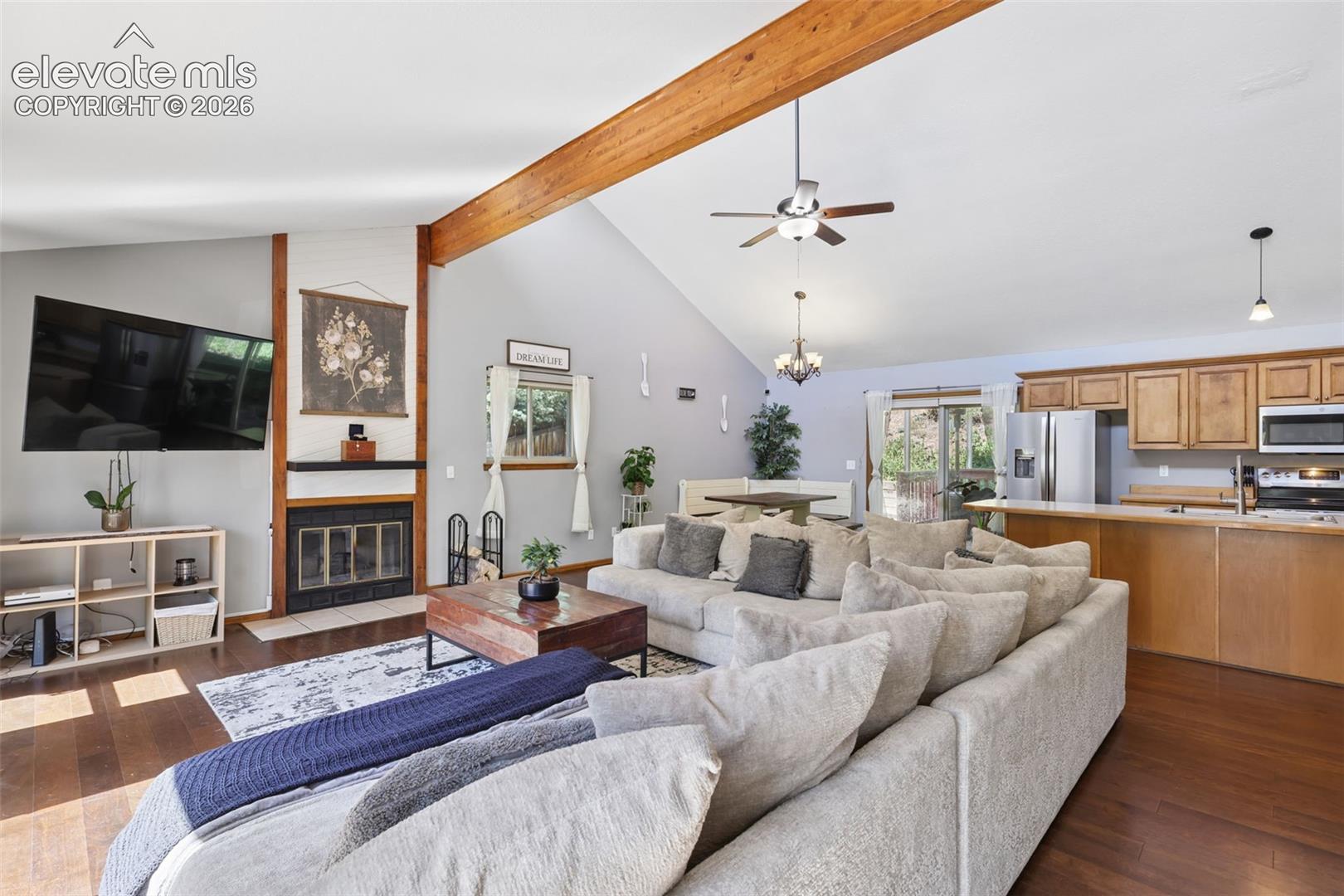Vaulted ceilings with exposed wood beams, a fireplace with a black tile surround, wood-finish flooring, a ceiling fan, and built-in kitchen cabinetry with stainless steel appliances