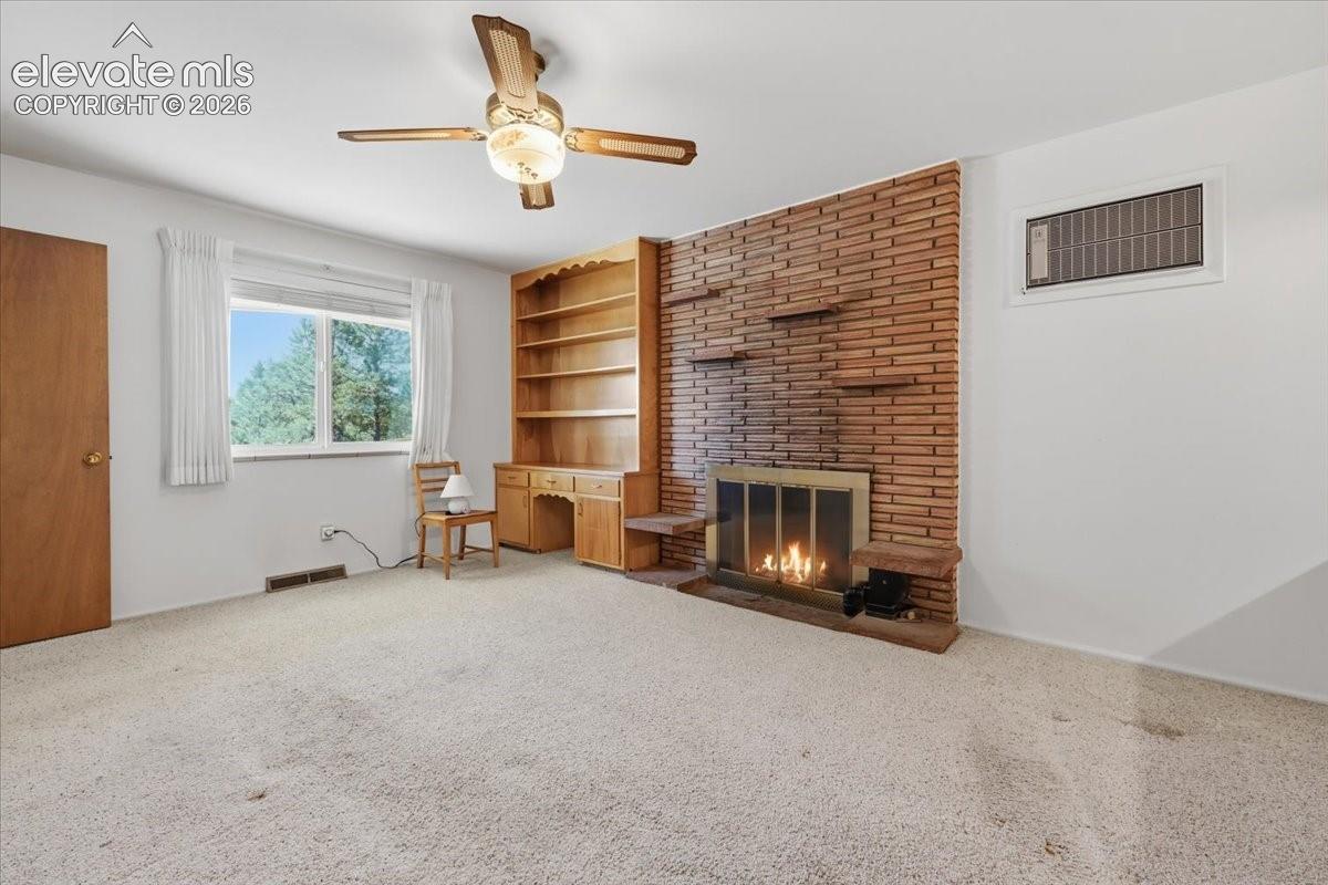 Floor-to-ceiling brick fireplace with attached shelving, built-in wooden cabinetry with desk workspace, carpeted flooring, and a multi-blade ceiling fan with integrated lighting