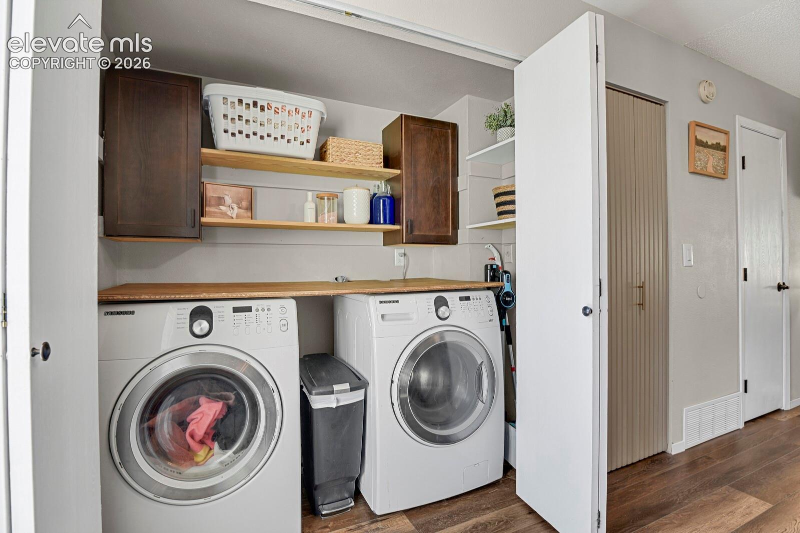 Laundry area featuring wood-finish flooring, two front-loading appliances, a light wood-finish countertop, and wall-mounted dark wood-finish cabinetry