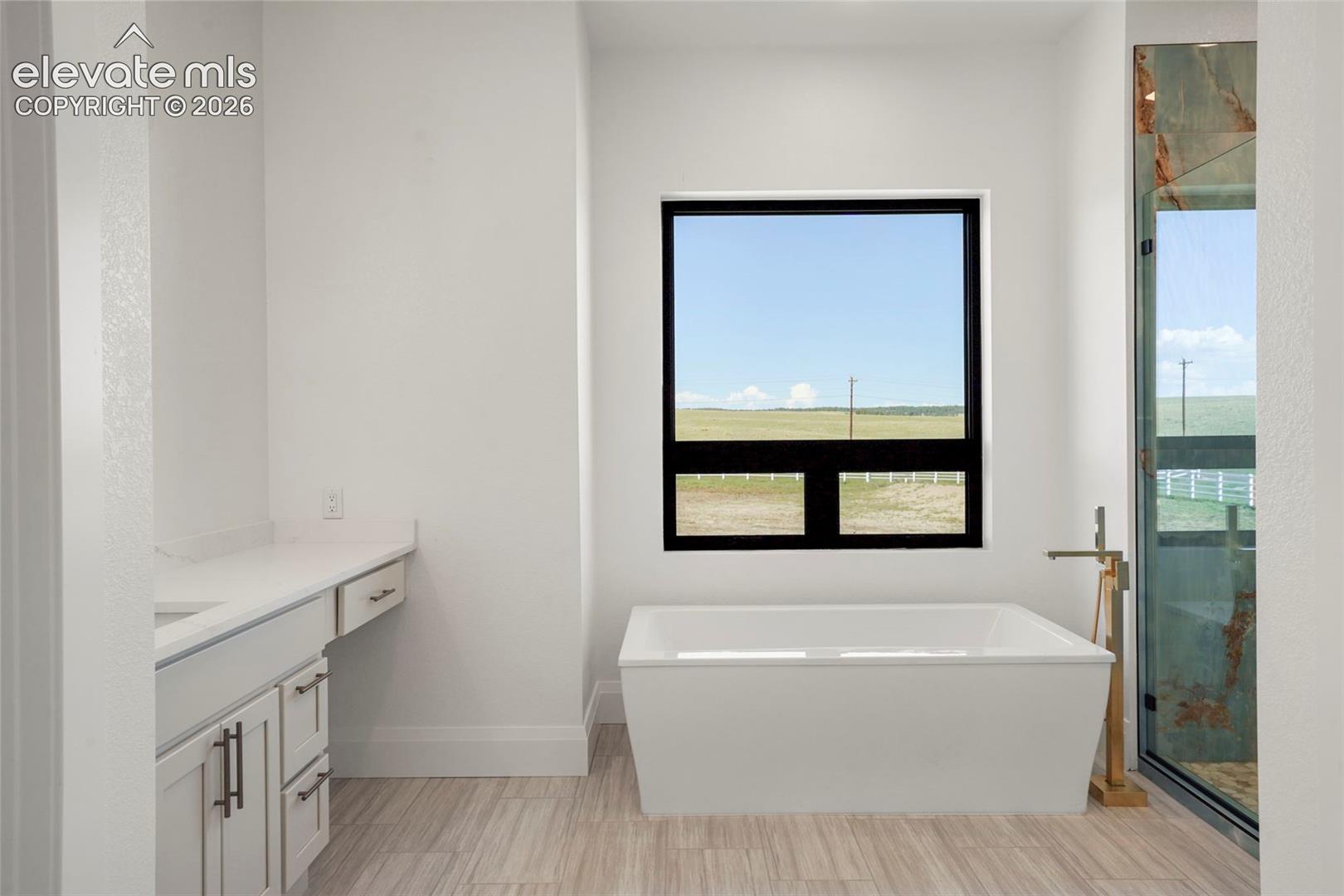Freestanding soaking tub with a floor-mounted gold-tone faucet, large black-framed window, white shaker-style cabinetry with dark hardware, and light-toned wood-finish flooring