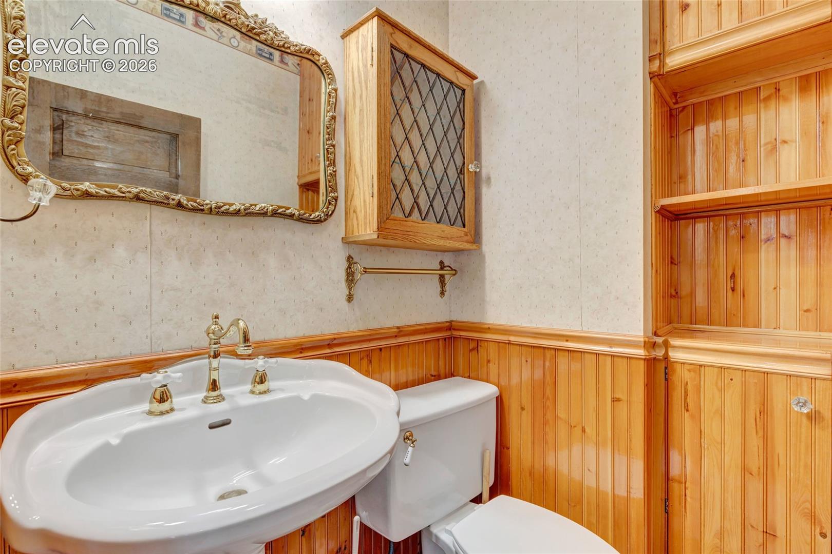 Bathroom featuring wood-finish wainscoting, built-in shelving, wall-mounted cabinet with leaded glass inlay, and a pedestal sink with brass-finish hardware