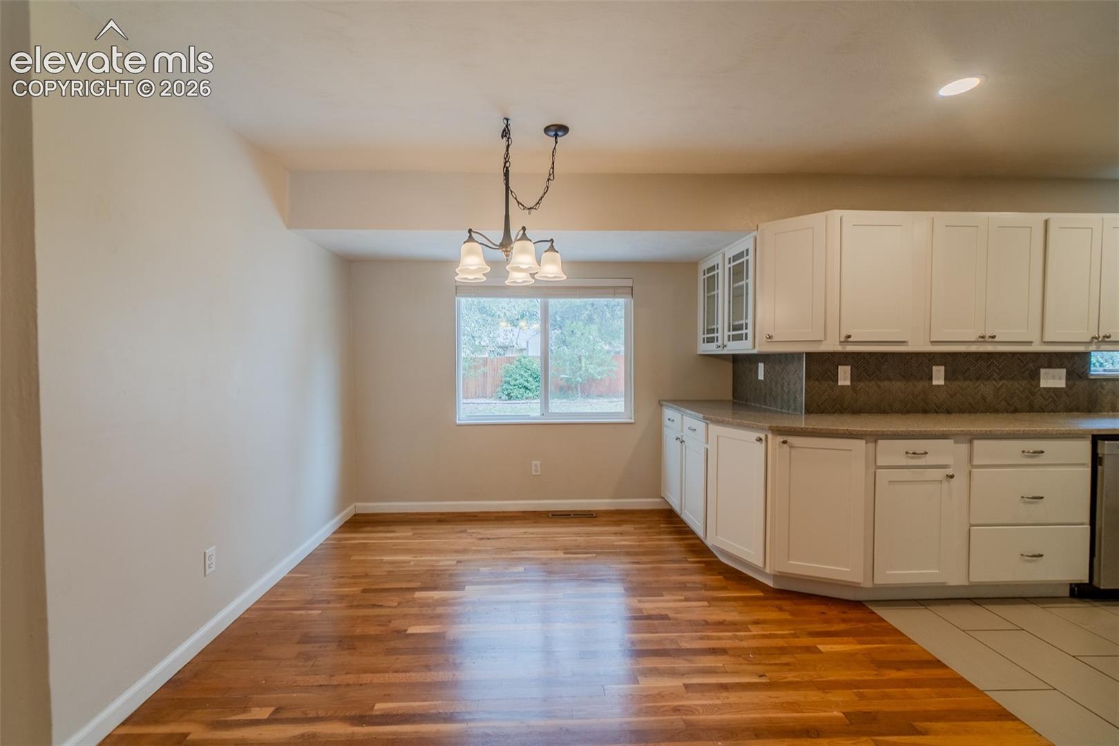 Wood-finish flooring, white cabinetry, dark countertop, tile backsplash, and a multi-bulb chandelier