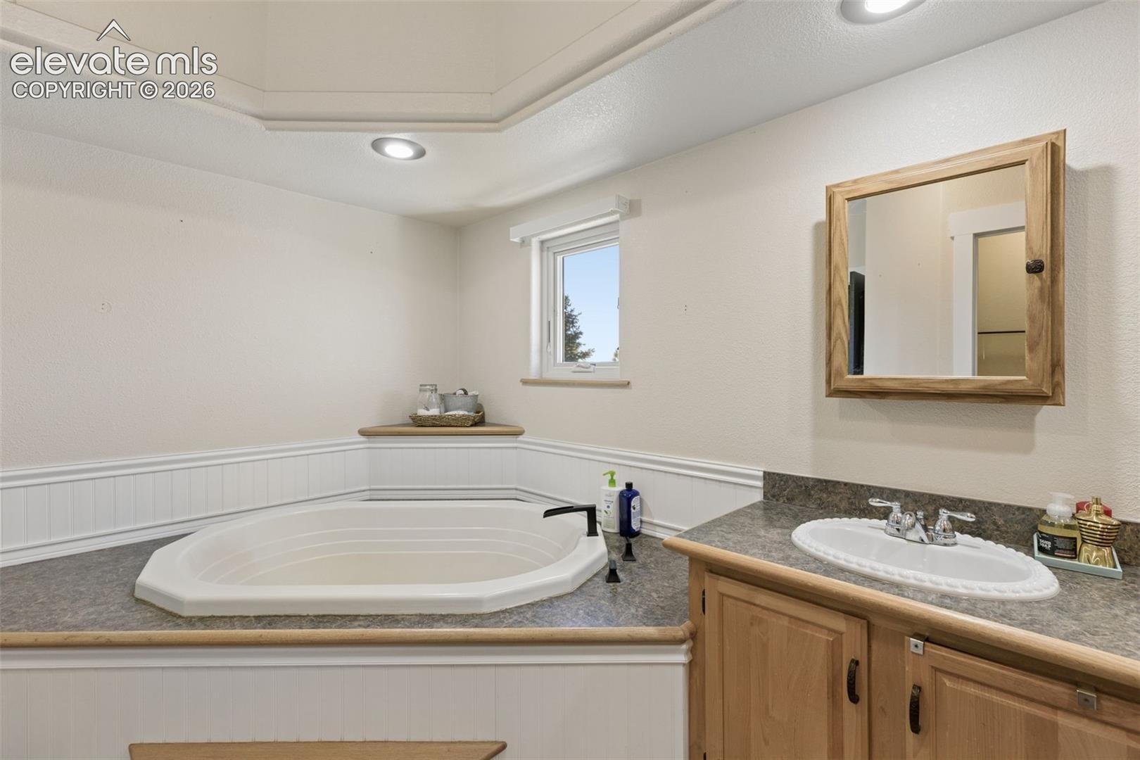 Built-in jetted soaking tub with tile surround, wood-finish cabinetry, drop-in sink with metal hardware, wall-mounted medicine cabinet, and recessed ceiling lighting