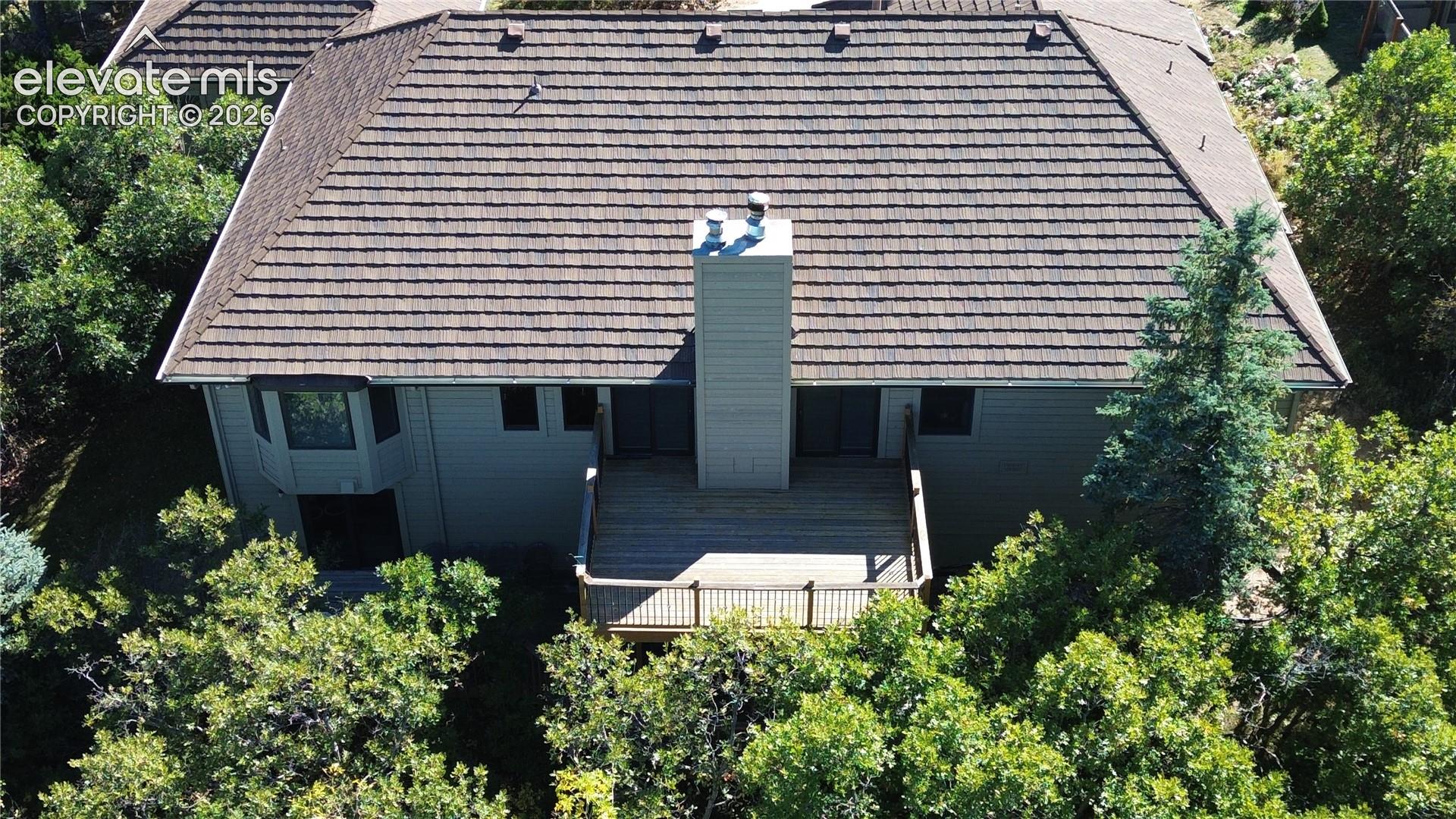 Aerial perspective featuring a multi-level wood deck, central chimney, shingled hip roof, and surrounding mature trees