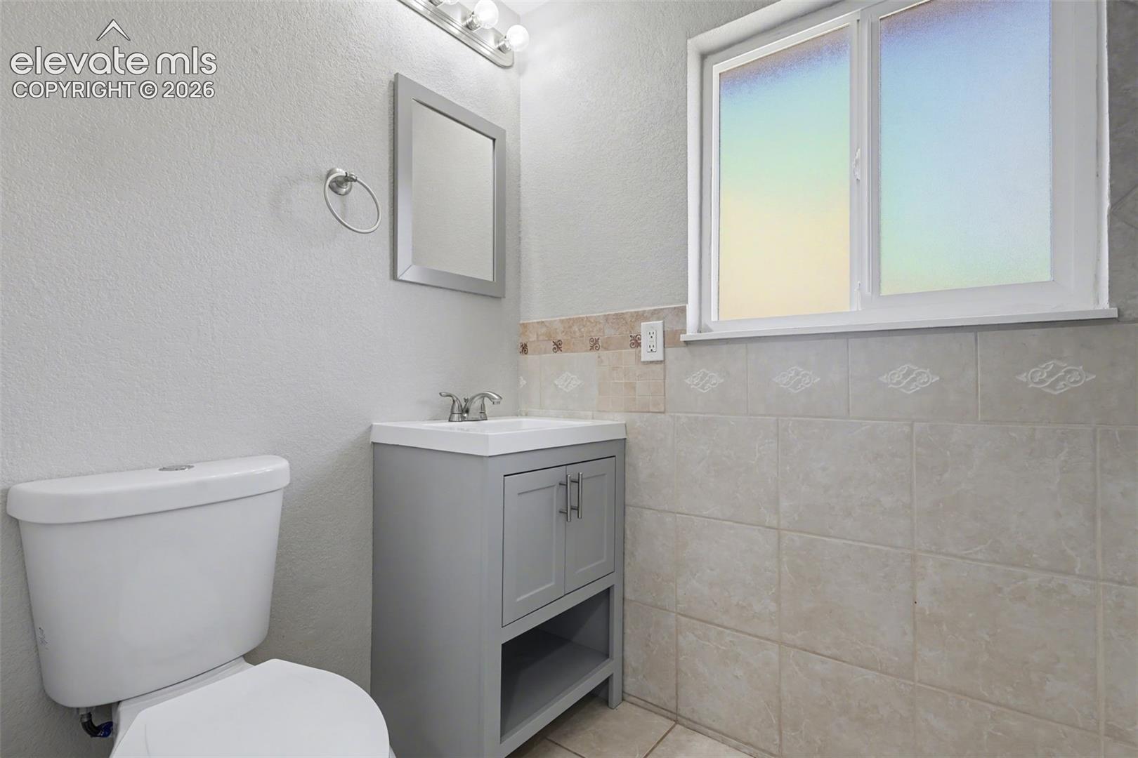Gray vanity with cabinet storage, porcelain-surfaced sink, dual-handle faucet, neutral tile wainscoting, and rectangular window with frosted privacy glass