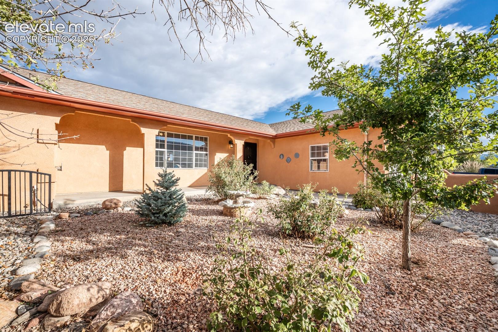 Stucco exterior featuring a covered entryway, decorative corbels, xeriscaped front yard with river rock ground cover, and metal gate
