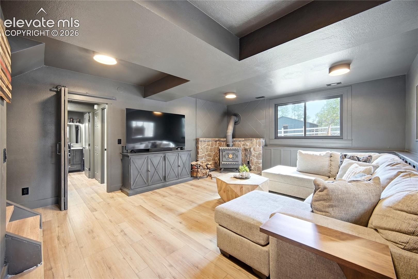 Living space featuring wood-finish flooring, a stone-surround wood stove, exposed ceiling beams, a sliding barn door, and a built-in media console