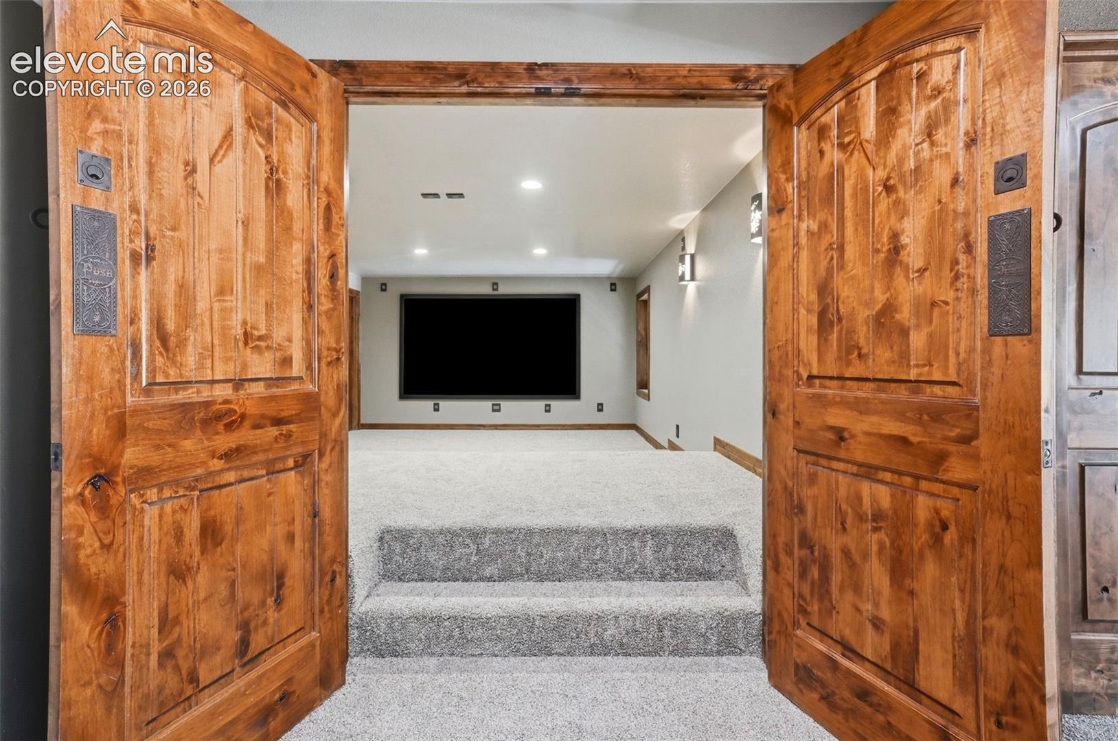 Double wood doors with dark hardware open to a carpeted room featuring a large screen, recessed lighting, and wall sconces