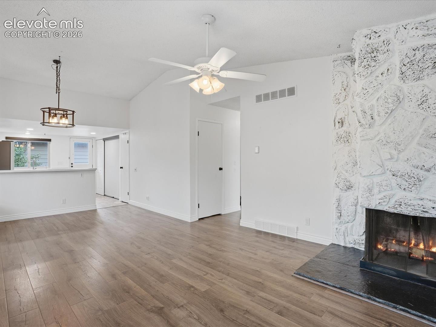 Living area featuring vaulted ceilings, wood-finish flooring, a full-height stone-clad fireplace, and a hanging pendant light fixture