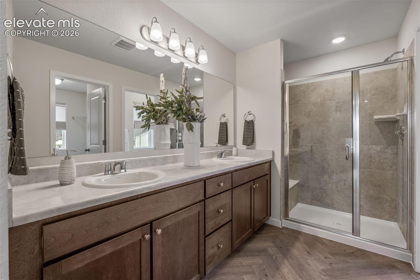 Dual vanity with white countertop and wood-finish cabinetry, frameless mirror, and glass-enclosed shower with tiled walls and built-in bench