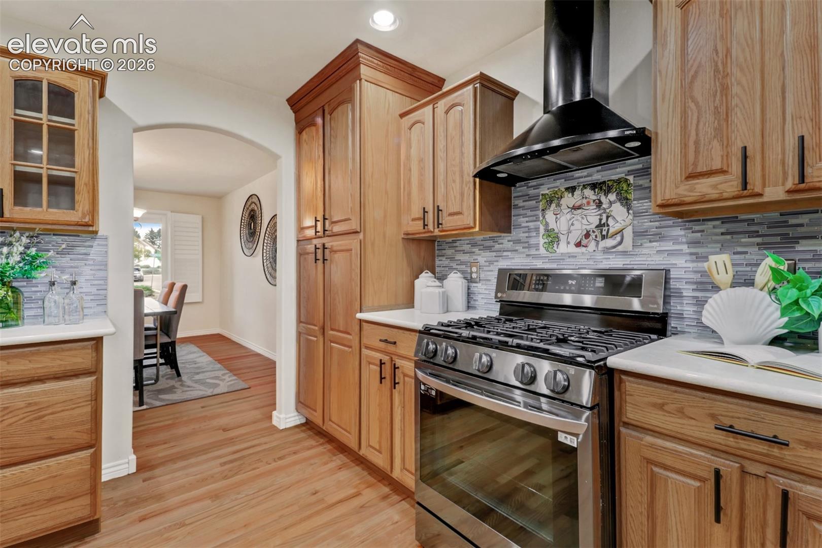 Kitchen featuring wood cabinetry, stainless steel gas range, black chimney-style range hood, solid-surface countertops, and glass tile backsplash