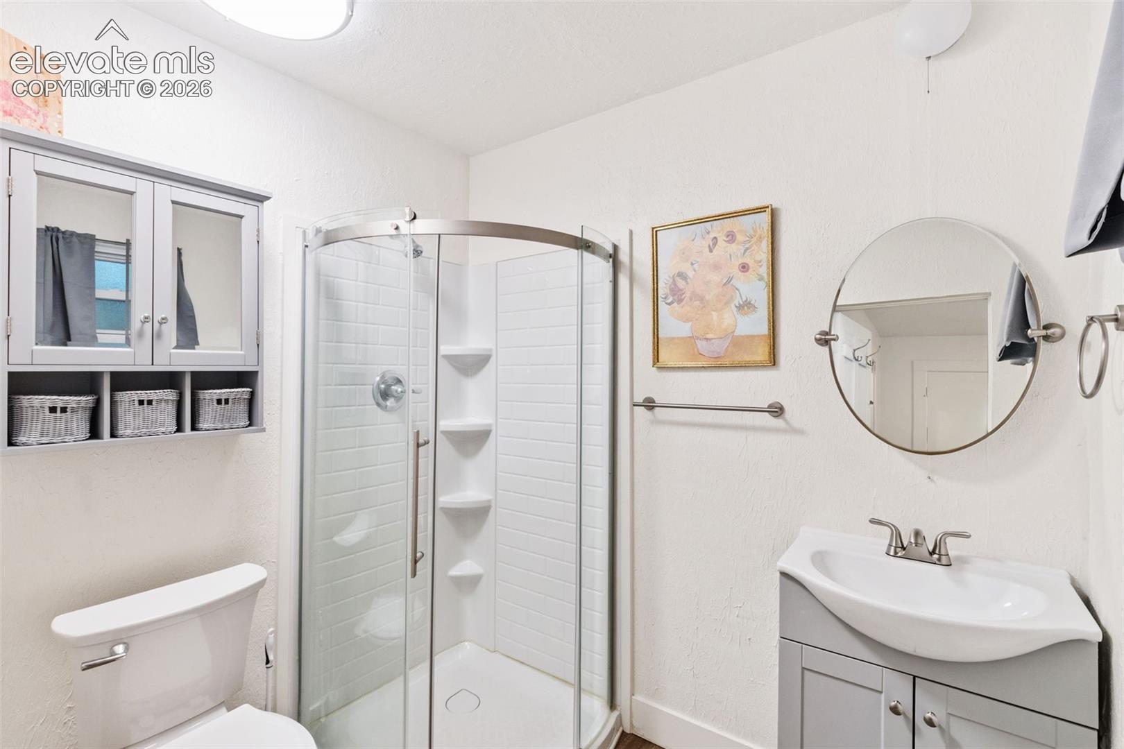 Bathroom featuring a corner shower with glass enclosure, grey vanity cabinet, pedestal-style sink, wall-mounted medicine cabinet with open shelving, and a circular mirror