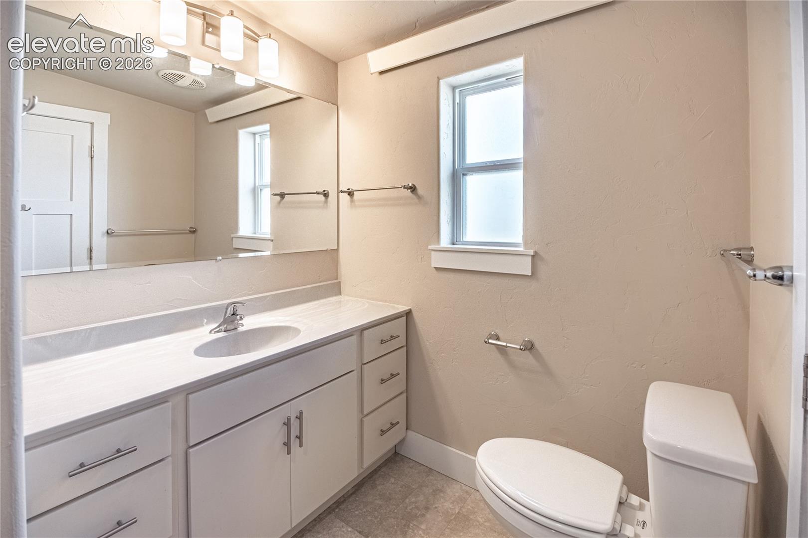 Bathroom featuring an elongated white toilet, a white vanity with an integrated sink and chrome faucet, a large mirror, and tile flooring