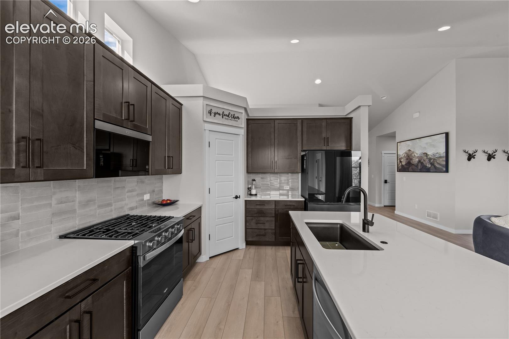 Kitchen featuring dark-toned cabinetry, solid-surface countertops, tile backsplash, wood-finish flooring, and a large center island with an undermount sink and matte black faucet