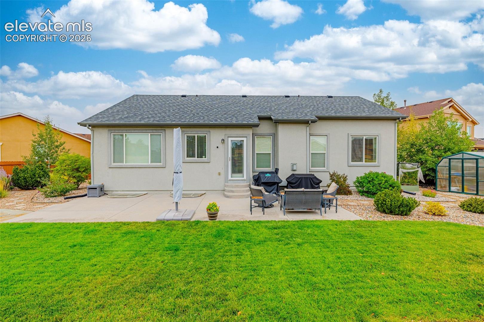 Rear exterior featuring a concrete patio, stucco siding, a grey shingle roof, manicured lawn, and low-maintenance gravel landscaping
