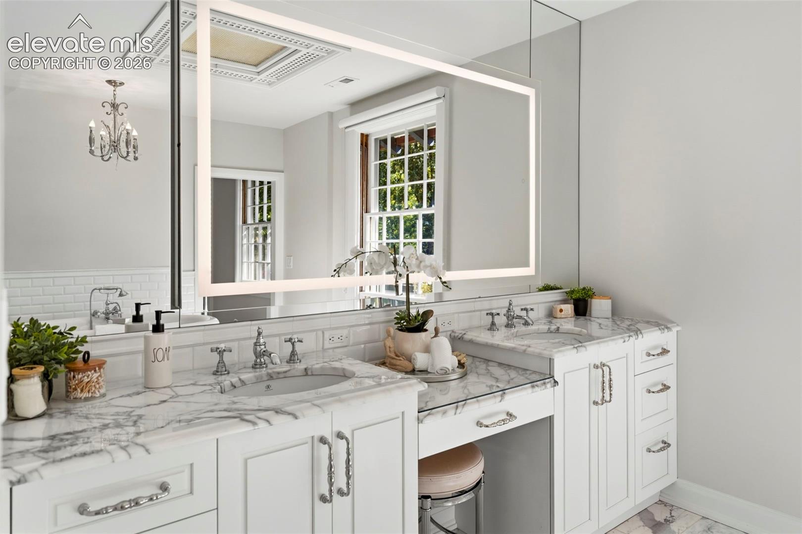 Double vanity with stone countertops, integrated oval basins, chrome hardware, backlit mirror, and white cabinetry