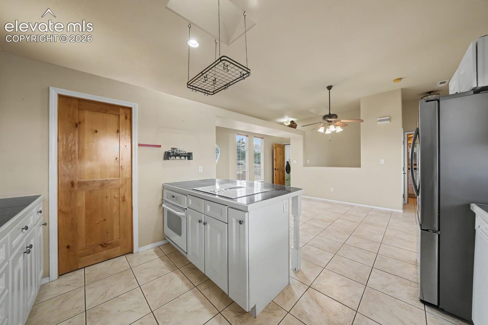 Kitchen featuring an island with a built-in cooktop, neutral tile flooring, a metal ceiling-mounted pot rack, solid wood door, and stainless steel refrigerator