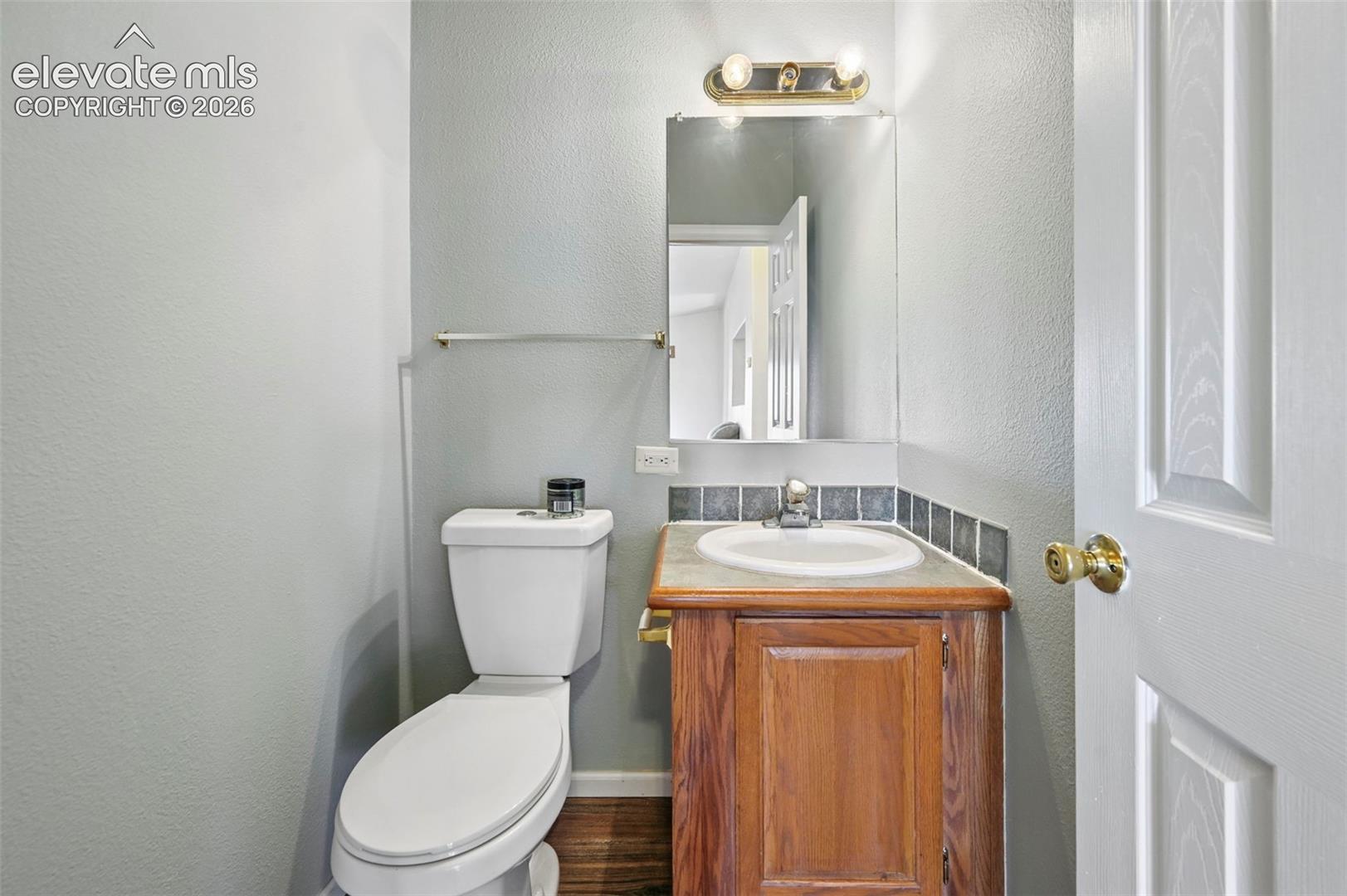 Restroom featuring a wood-finish vanity with a stone countertop, integrated sink, tiled backsplash, wall-mounted mirror, and metallic light fixture