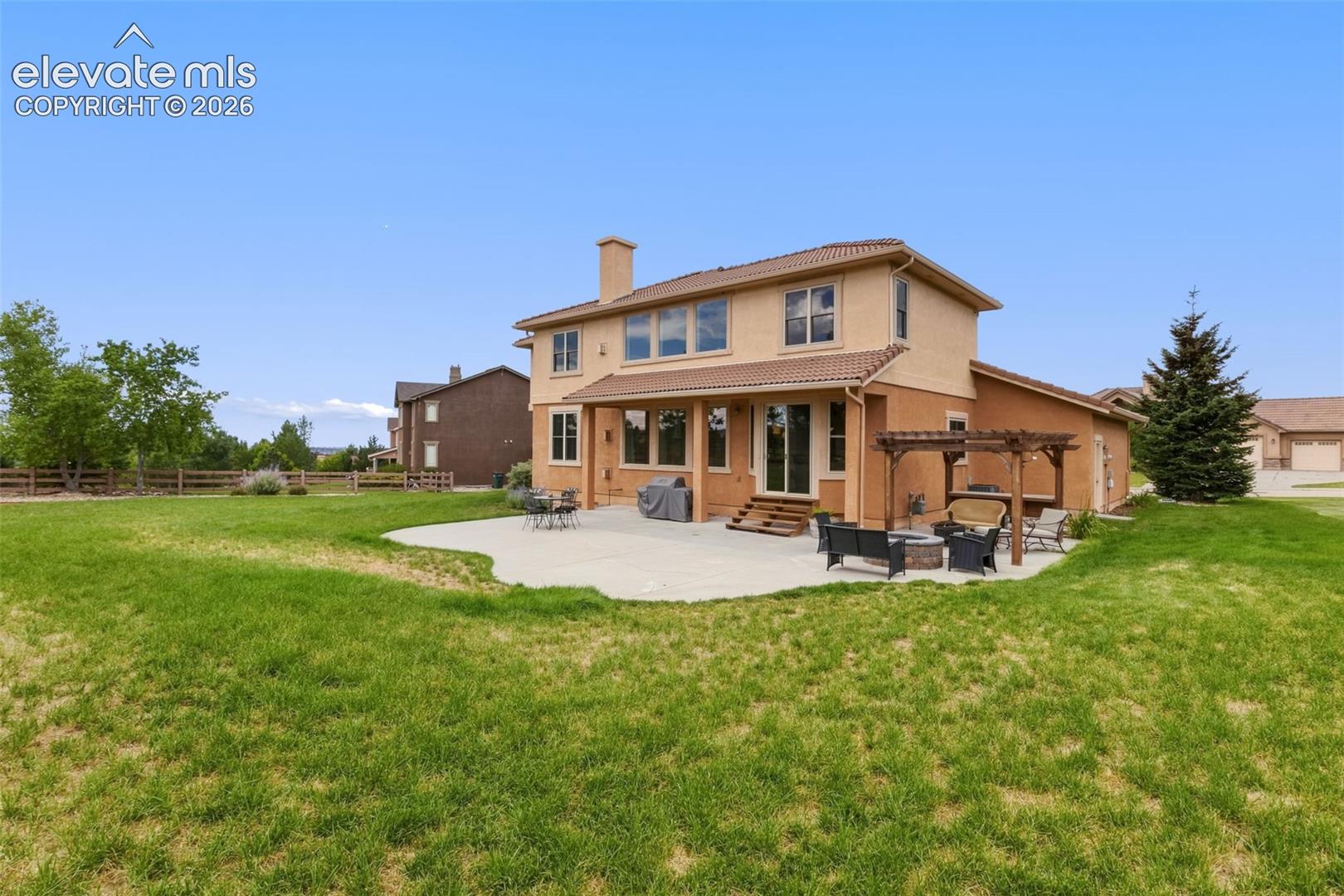 Expansive backyard featuring a large concrete patio with a wooden pergola, stucco exterior, and a tiled roof