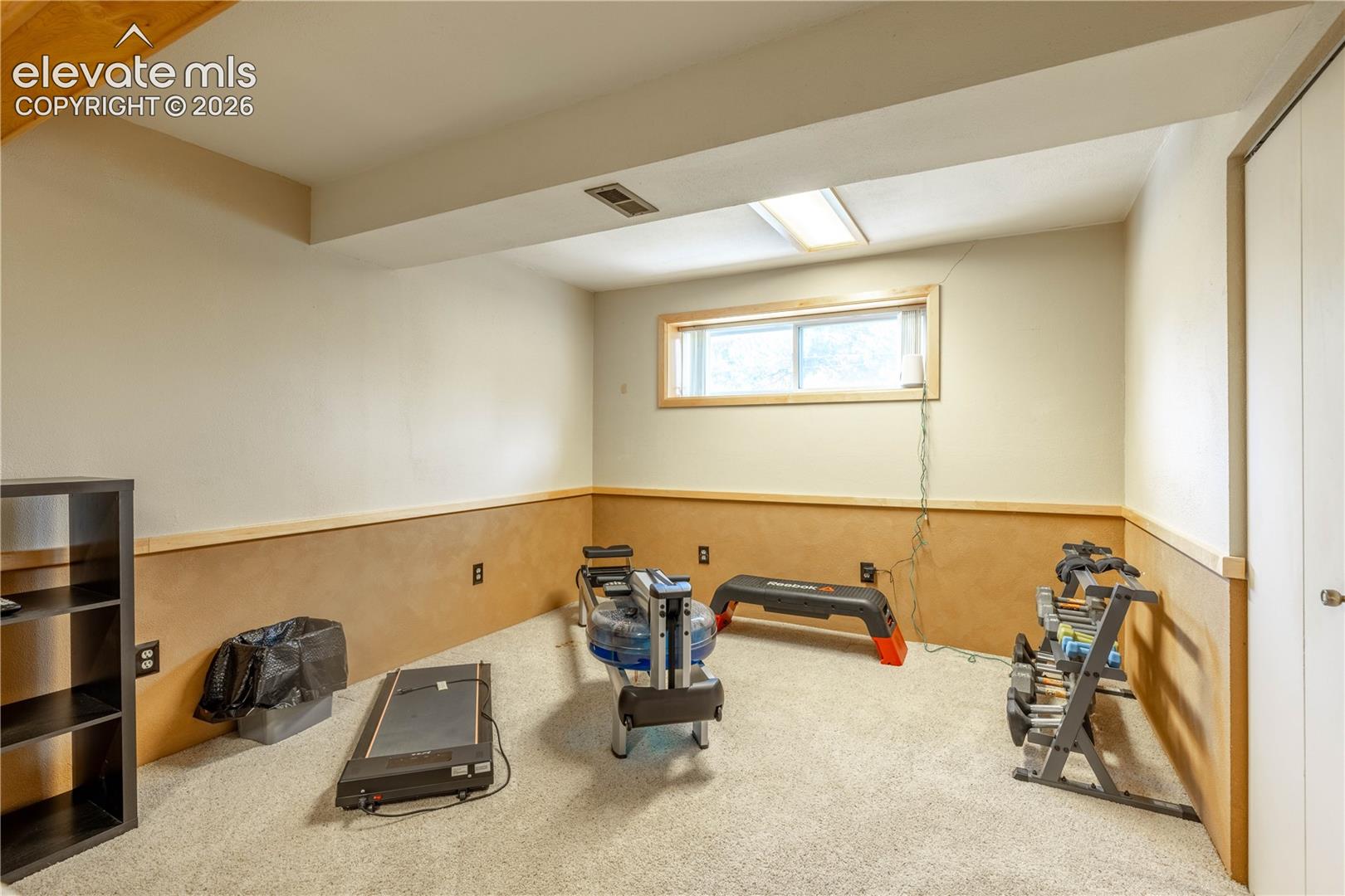 Neutral-toned interior space featuring light-colored wall-to-wall carpeting, wood-paneled lower wall accents, a rectangular window with wood casing, and a recessed ceiling light fixture