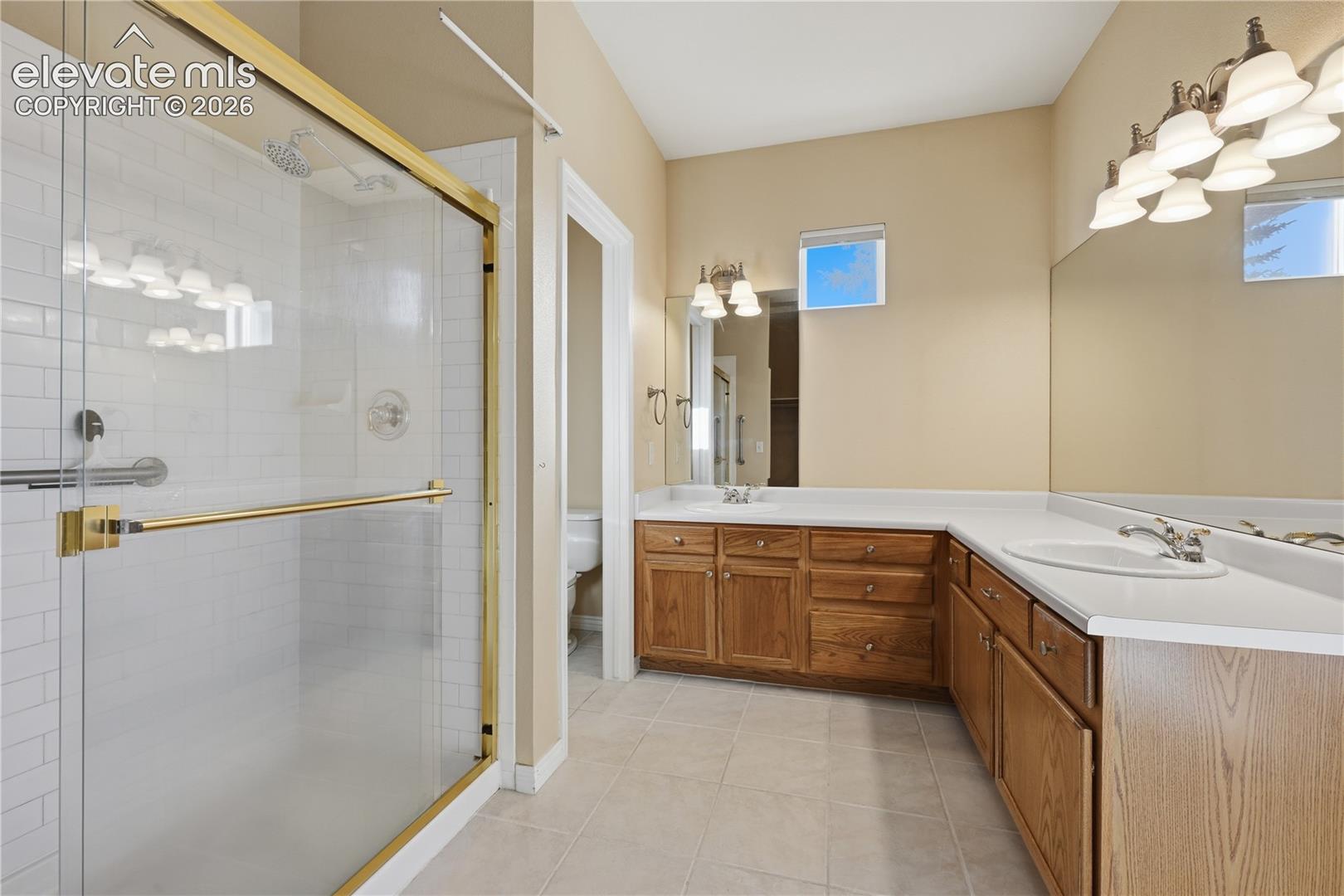 Bathroom featuring a dual-sink vanity with wood cabinetry, a solid surface countertop, a walk-in shower with white subway tile surround and gold-tone frame, and tile flooring