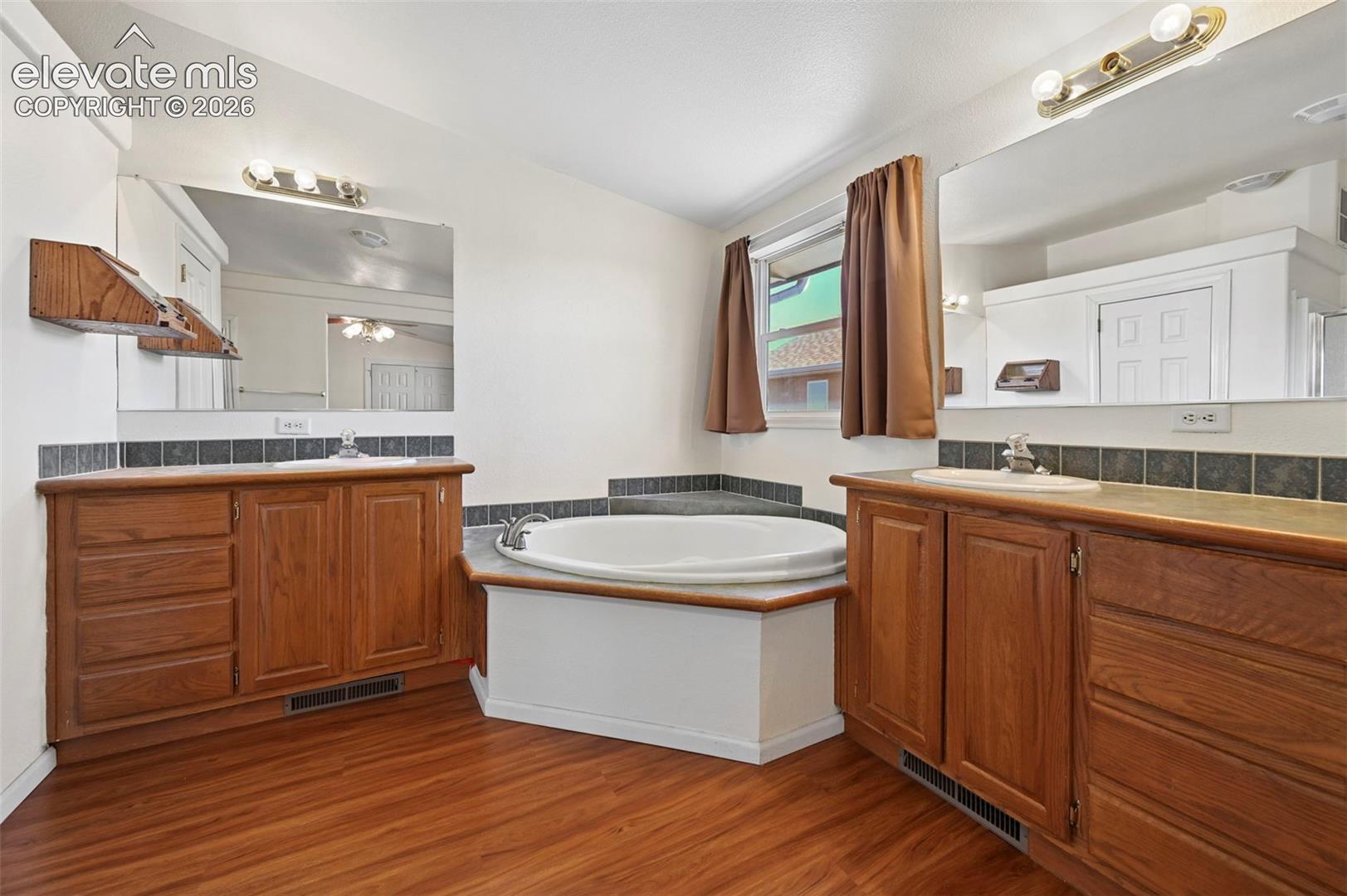 Dual vanities with wood cabinetry, drop-in sinks, tiled backsplash, large wall-mounted mirrors, and a central jetted soaking tub