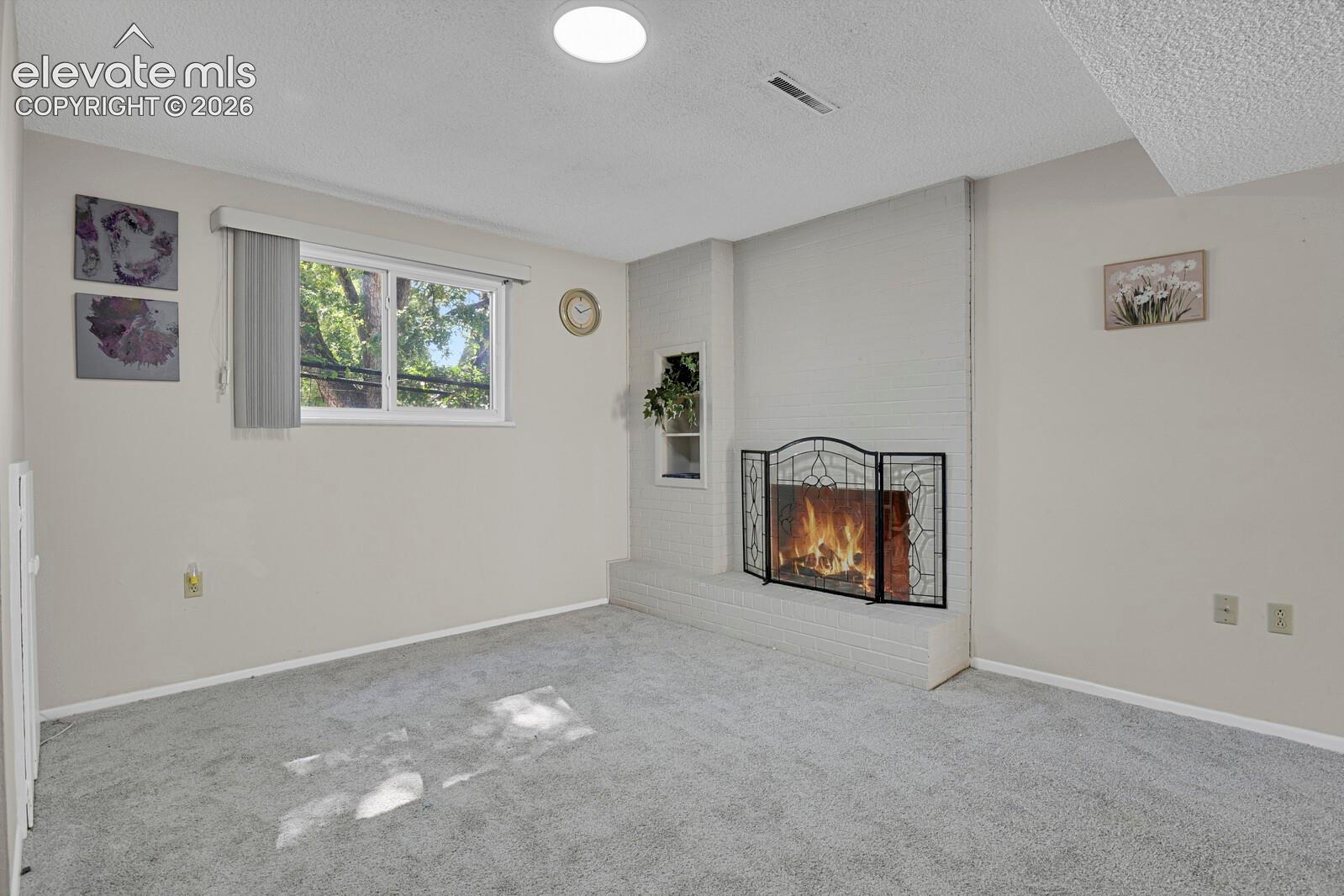 Carpeted interior space featuring a white brick fireplace with a black metal screen, a recessed shelf, and a window with a white frame and vertical blinds