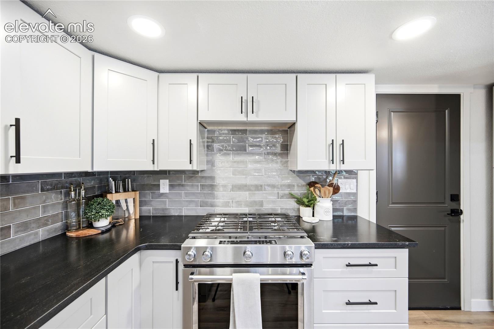 White shaker-style cabinetry with matte black hardware, dark countertops, subway-patterned tile backsplash, and stainless steel gas range