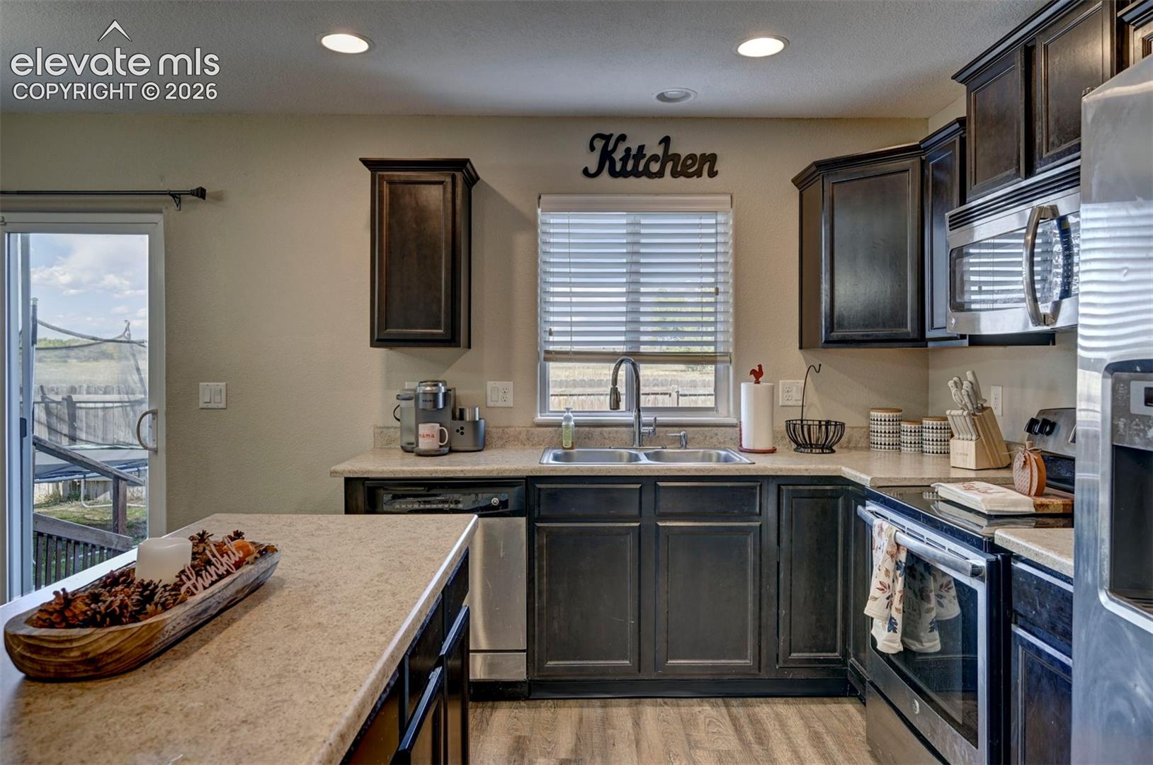 Kitchen featuring dark wood-finish cabinetry, stainless steel appliances, a double basin sink with a high-arch faucet, and recessed lighting