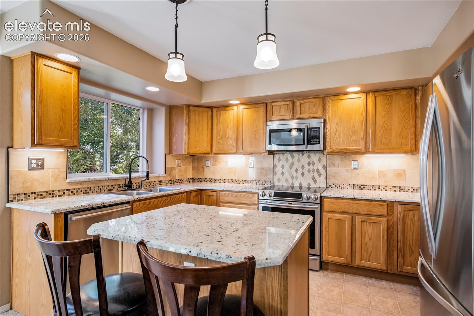 Warm wood cabinetry, granite countertops, stainless steel appliances + a custom tile backsplash complement the kitchen's pendant lighting and convenient center island.