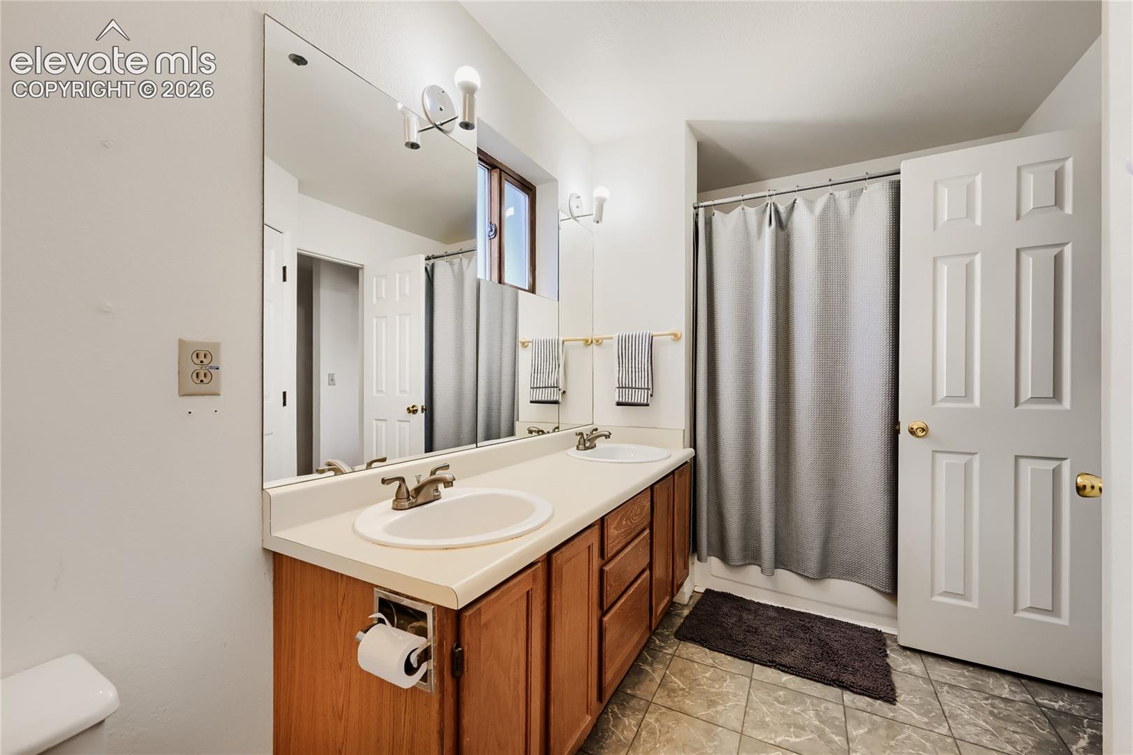 Spa-inspired bathroom featuring a wood-finish vanity with dual-sink configuration, ceramic countertop, and brushed bronze faucetry