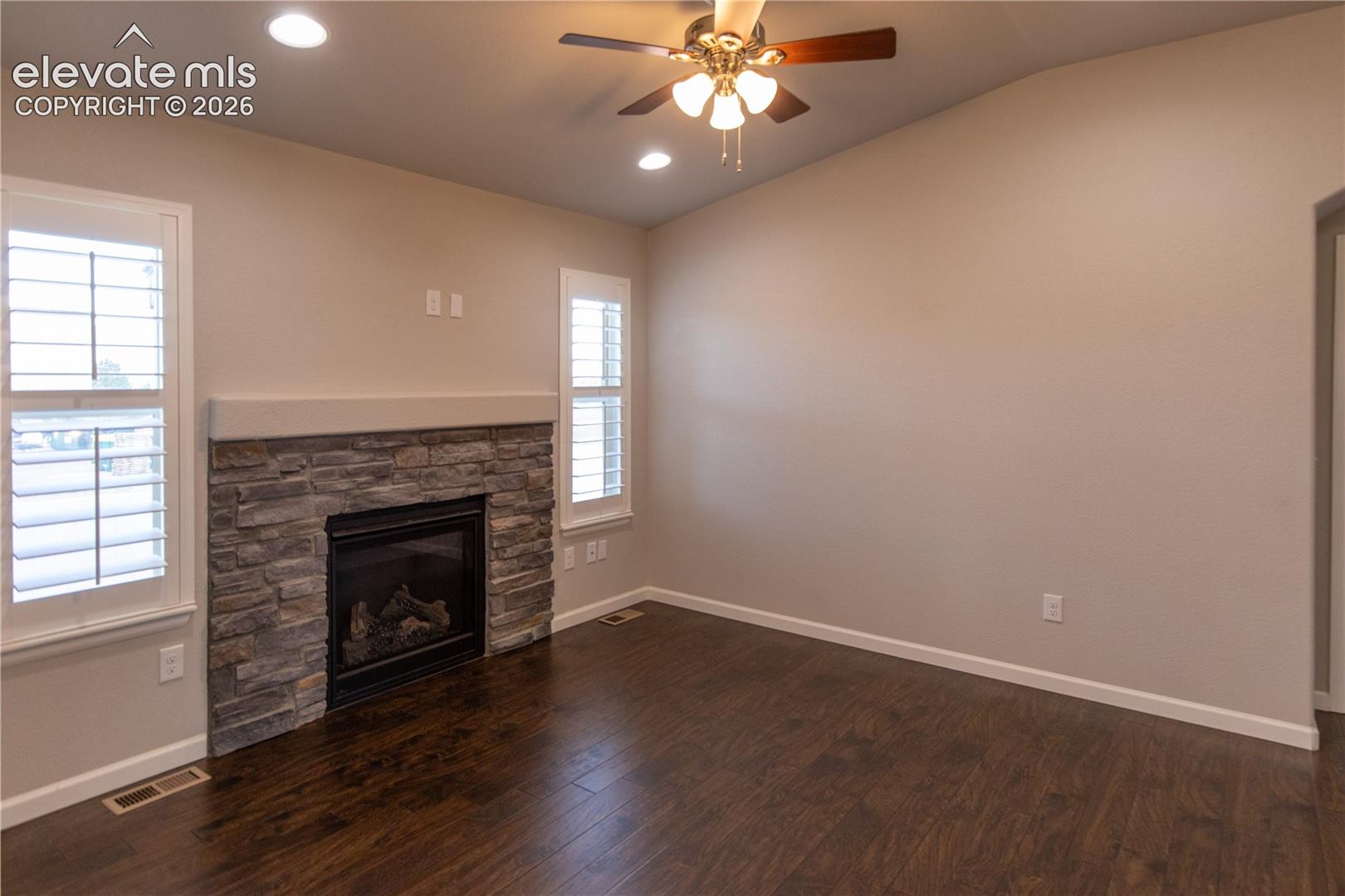 Living area featuring wood-finish flooring, stone-veneer fireplace with a mantle, vaulted ceiling, recessed lighting, and interior shutters