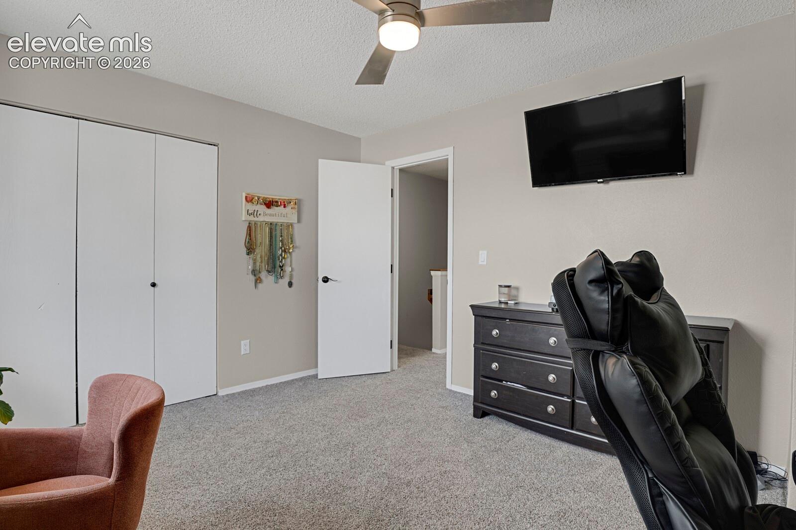 Textured carpeting, neutral wall paint, ceiling fan, white bi-fold closet doors, and a wall-mounted television