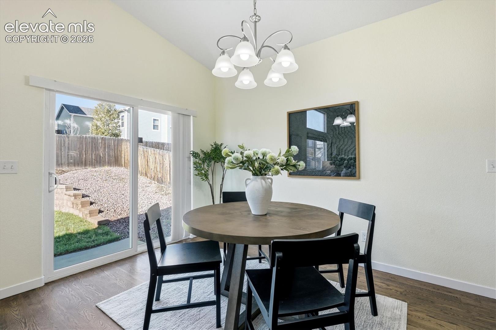 Vaulted ceiling with a central chandelier, wood-finish flooring, and sliding glass door leading to a backyard