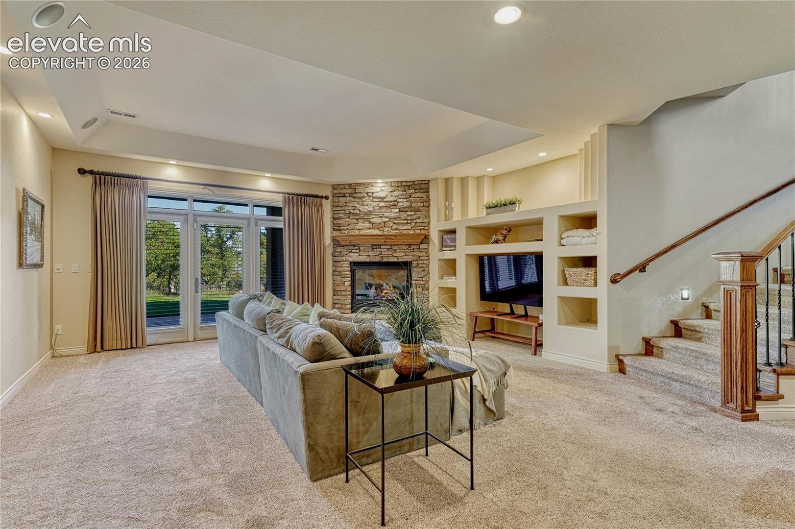 Corner fireplace featuring stacked stone surround and timber mantel, custom built-in shelving, tray ceiling with recessed lighting, and double glass doors