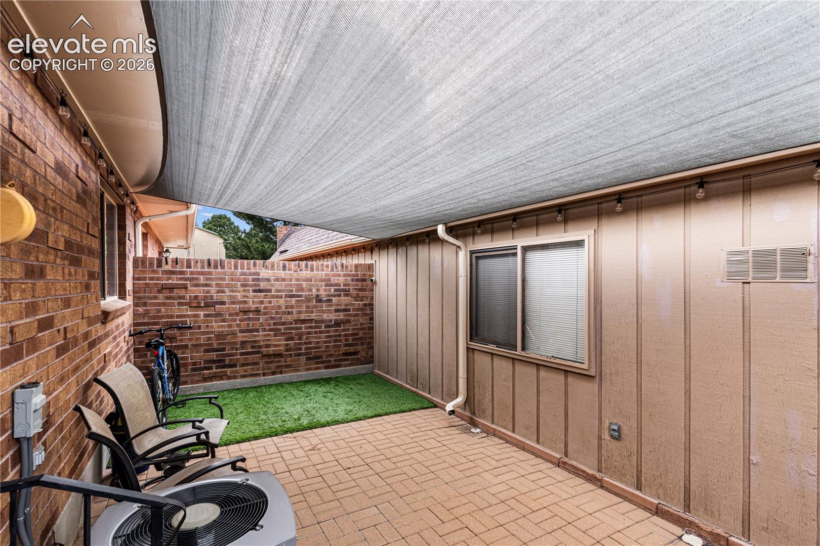 Private patio featuring a herringbone-patterned paver surface, brick masonry walls, a retractable fabric shade canopy, and synthetic turf lawn area