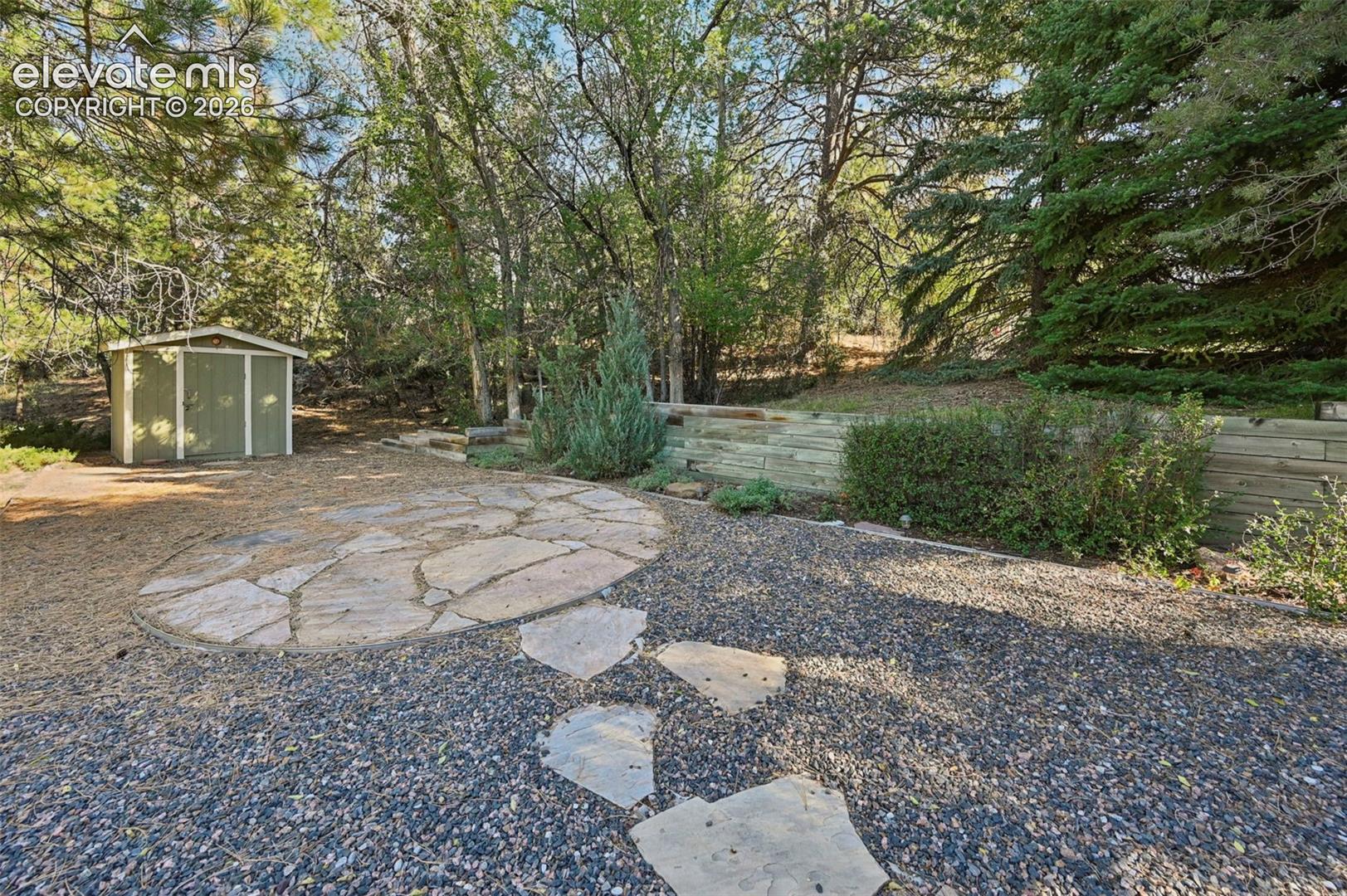 Backyard featuring a circular stone patio, gravel surfacing, timber retaining wall, and a detached storage shed set against a backdrop of mature trees