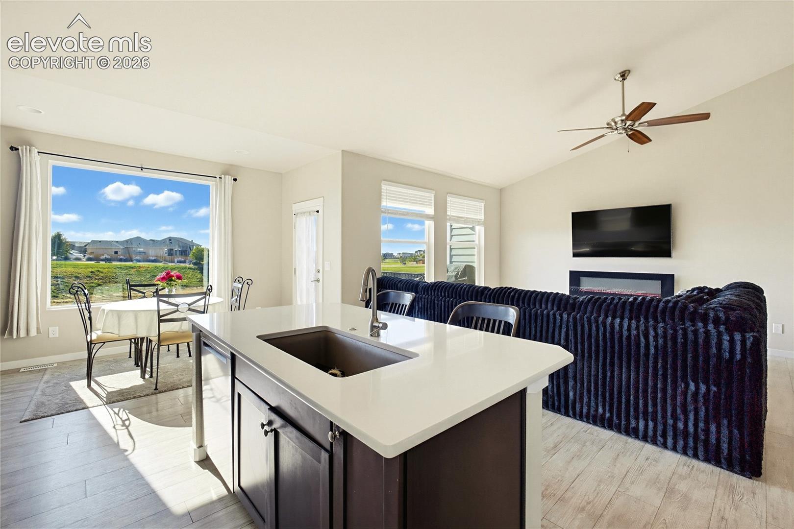 Kitchen island featuring a quartz countertop and integrated sink, vaulted ceilings, wood-finish flooring, wall-mounted electric fireplace, and expansive windows